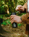 Person holding a black coffee grinder outdoors with greenery in the background