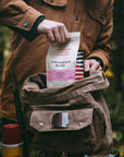 A person pulling a bag of Strongback Blend Coffee from their camping backpack outdoors in a forest.