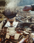 A person pouring boiling water into a Hario V60 maker while outdoors camping in the morning light