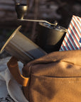 A backpack sitting outdoors with an Aeropress Brewer, Grinder and bag of coffee sticking out of the backpack.