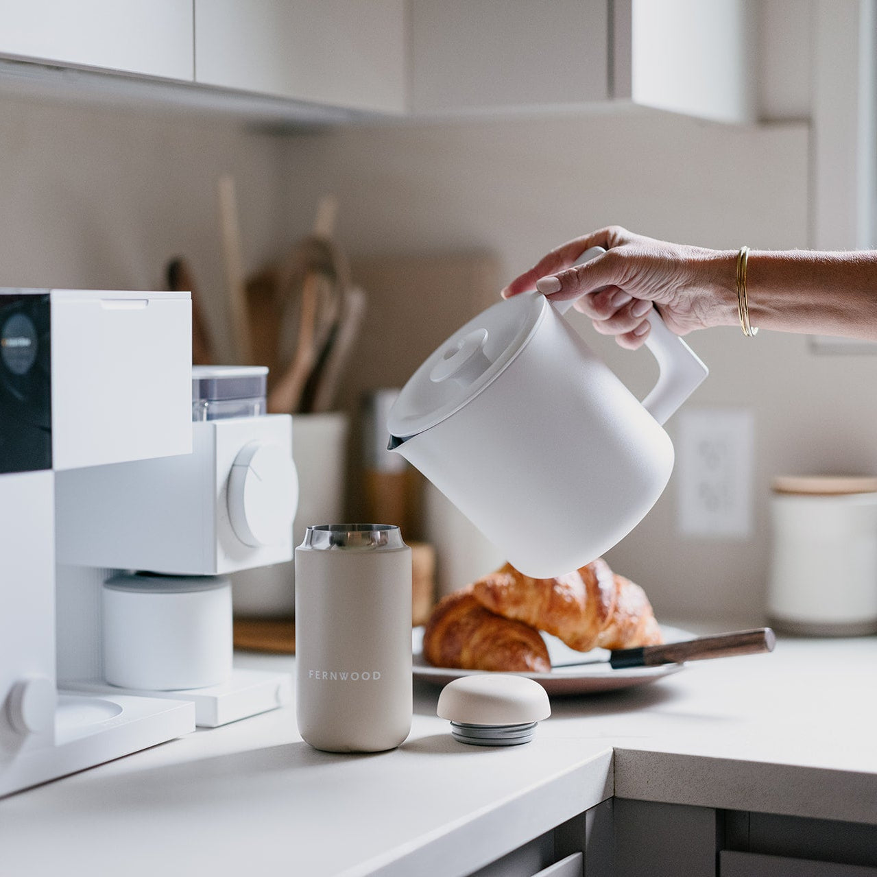 Person pouring from a white kettle into a mug on a kitchen counter with a microwave and croissant in the background.