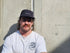 Man wearing a cap and white t-shirt with a logo against a concrete wall