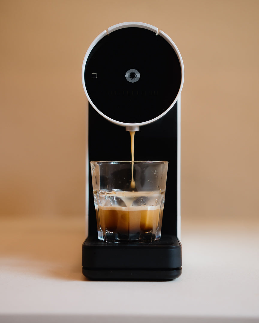 Capsule coffee machine dispensing coffee into a glass on a beige background