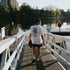 Person wearing a white tshirt walking on a wooden dock by a lake with buildings in the background