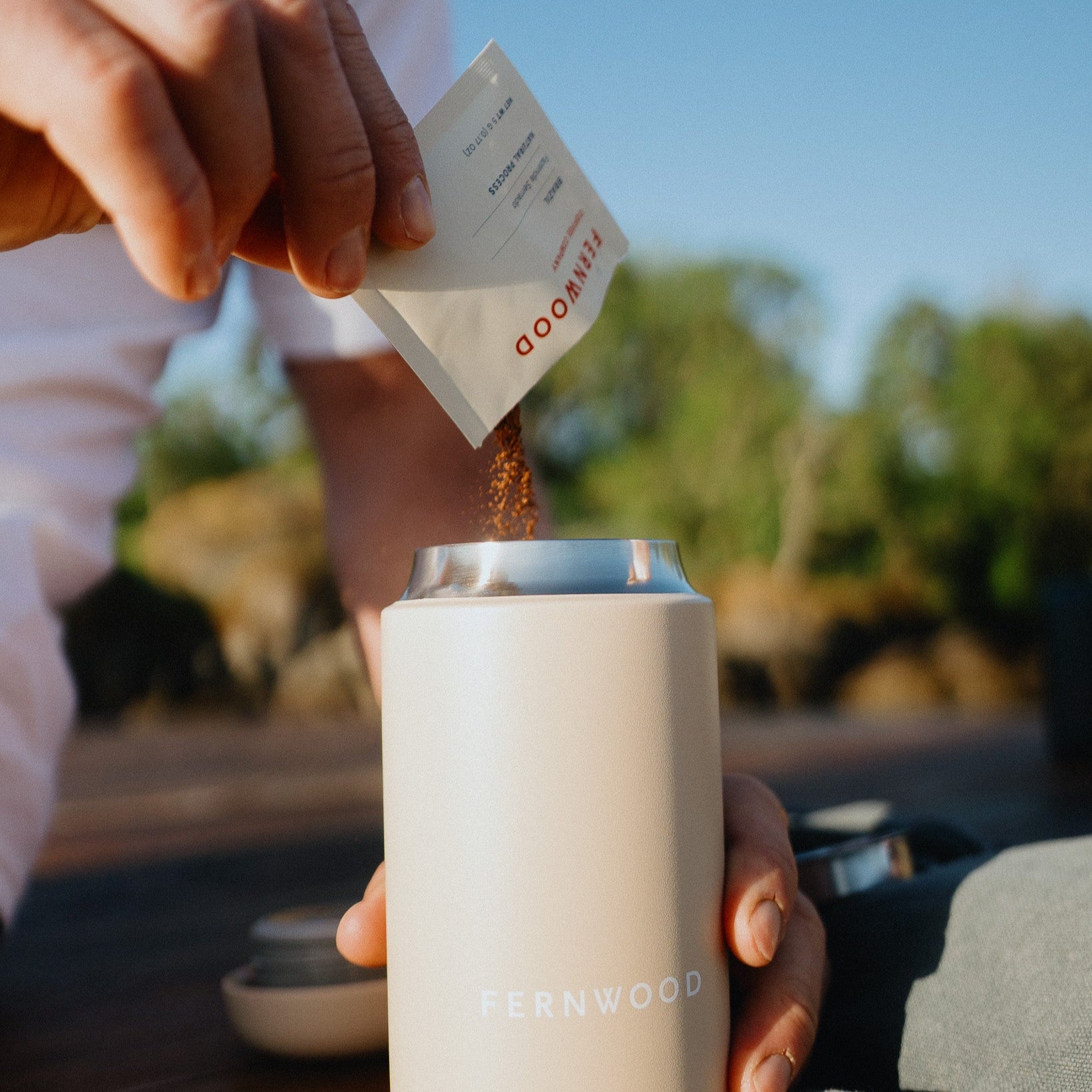 Person pouring instant coffee into a travel coffee tumbler with 'FERNWOOD' branding, outdoors.