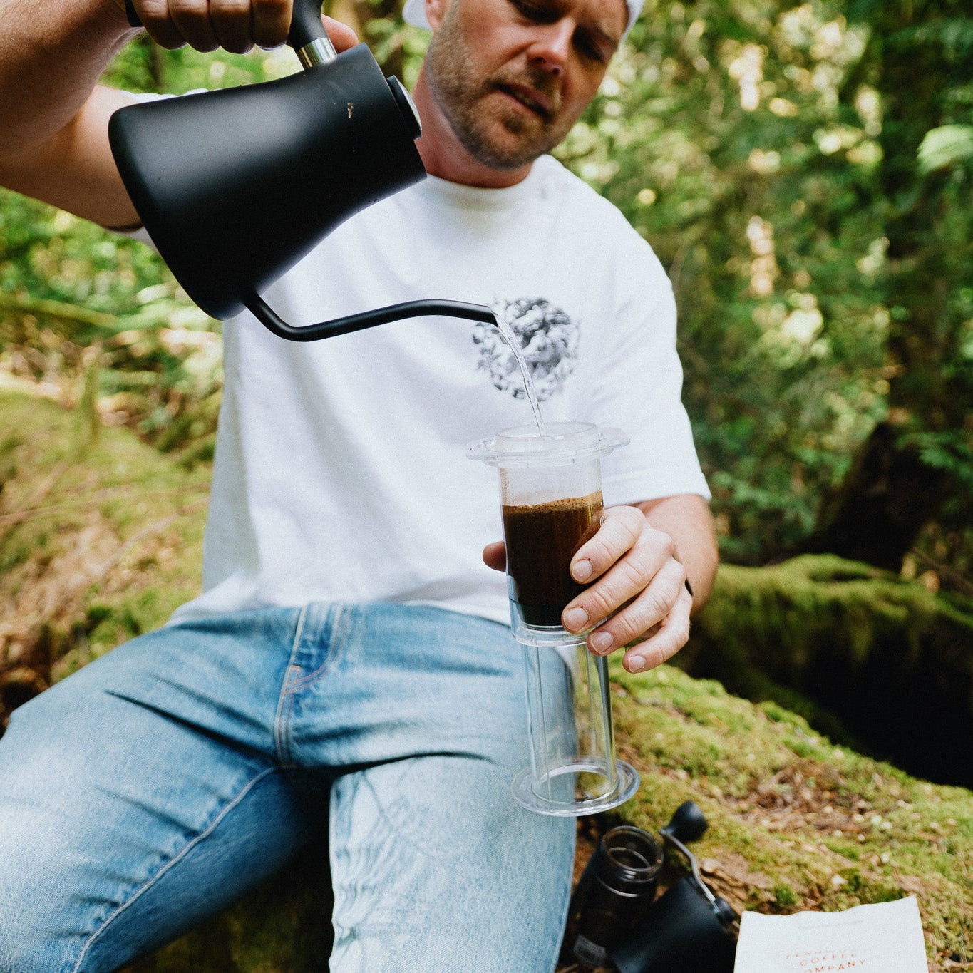 Man in a forest setting pouring coffee from a black coffee pot into a glass.