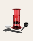 Red AeroPress coffee maker with black scoop and white filter on a beige background
