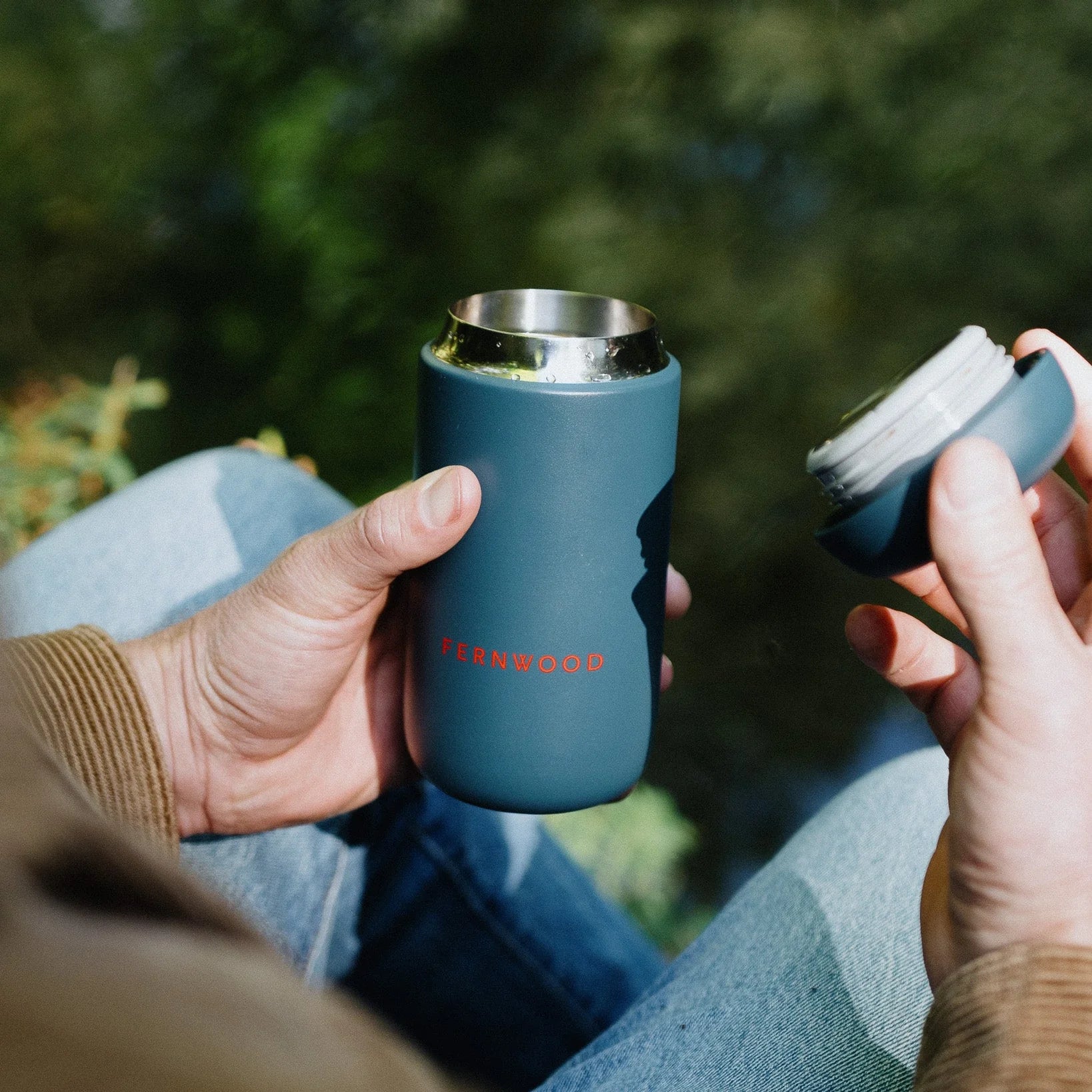 Person holding a blue travel mug with 'Fernwood' branding outdoors.