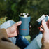 Person holding a blue travel mug with 'Fernwood' branding outdoors.
