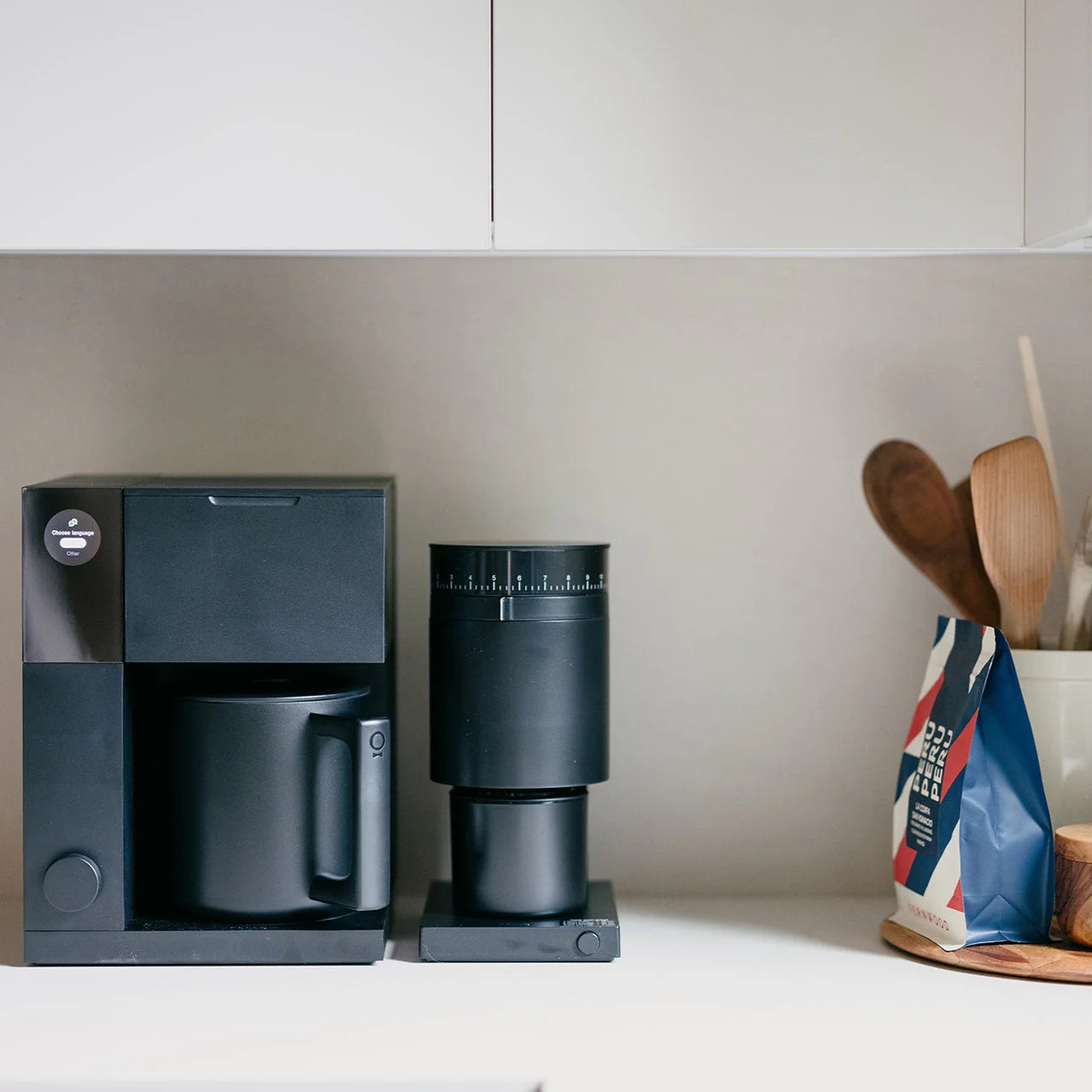 Black coffee maker and grinder on a kitchen counter with wooden spoons and a bag of coffee in the background.