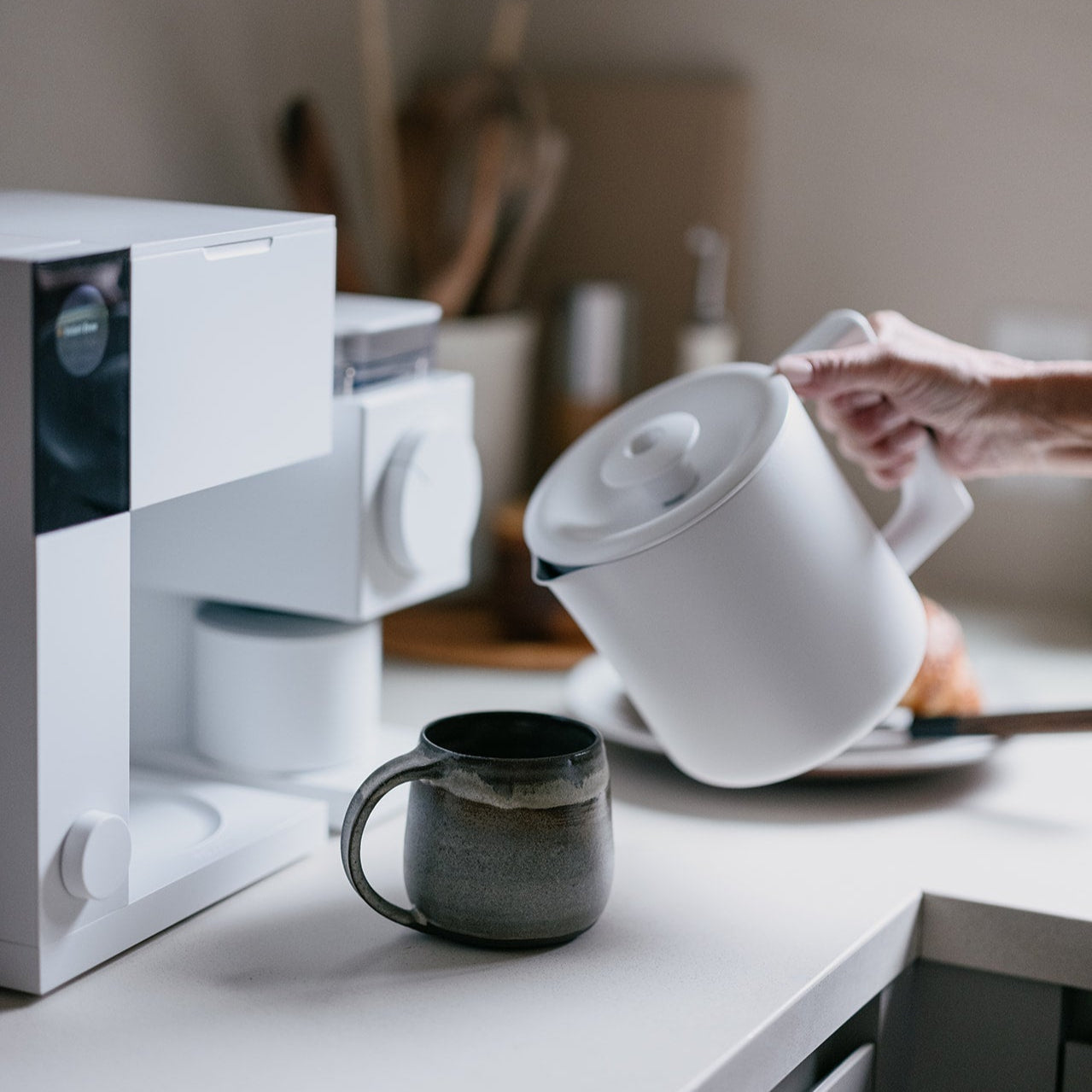 Person pouring coffee from a white coffee maker into a black mug on a kitchen counter.