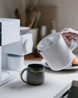 Person pouring coffee from a white coffee maker into a black mug on a kitchen counter.