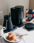 Black coffee maker on a kitchen counter with a croissant and coffee cup.
