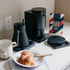 Black coffee maker, white mug, plate with pastry and knife on a kitchen counter.