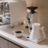 White kettle and coffee cup on a kitchen counter with a blurred coffee machine in the background.