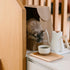 Cat peeking out from a cardboard box next to a kettle and cup of coffee on a wooden shelf.