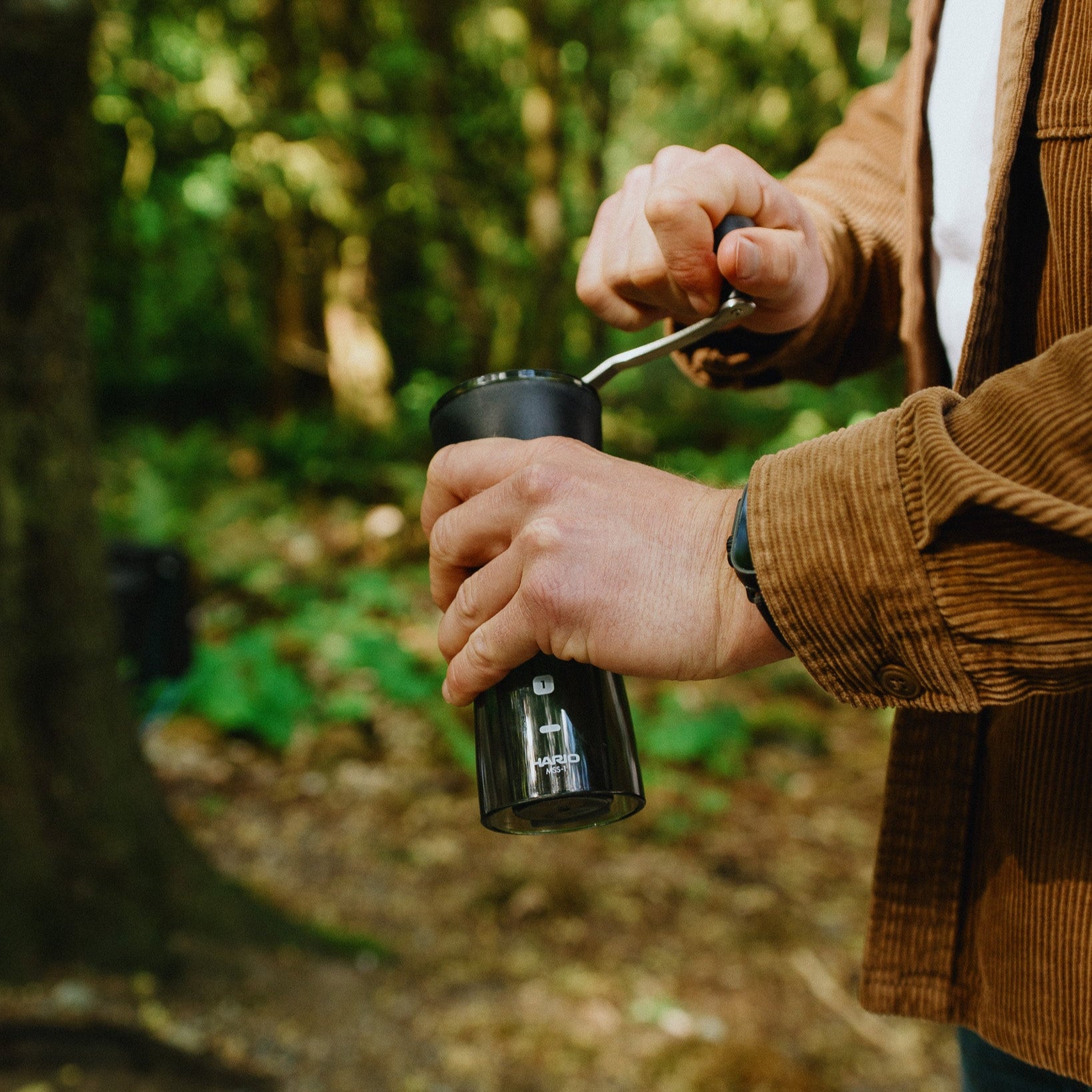 Person holding a black coffee grinder outdoors with greenery in the background