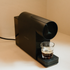 Black coffee machine dispensing coffee into a glass on a beige surface