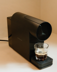 Black coffee machine dispensing coffee into a glass on a beige surface
