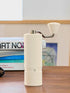 Off-white Nuttii Goosey hand grinder on a wooden table with books and coffee beans