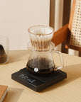 Coffee brewing setup with a glass carafe on a digital scale on a wooden table.