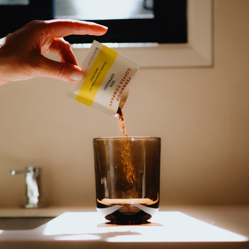 Hand pouring 1976 Espresso Instant coffee from a sachet into a glass on a kitchen counter.