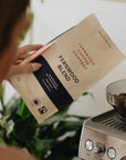 A person holding a bag of Fernwood Blend getting ready to pour beans into a grinder.