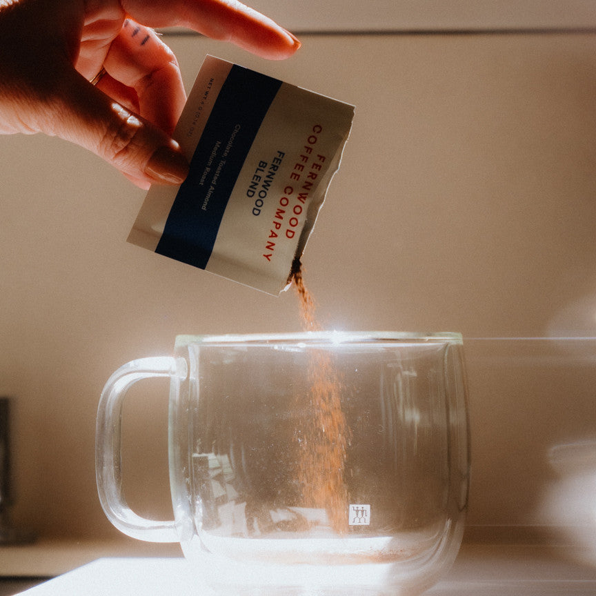 Hand pouring instant coffee from a pouch into a clear glass mug.