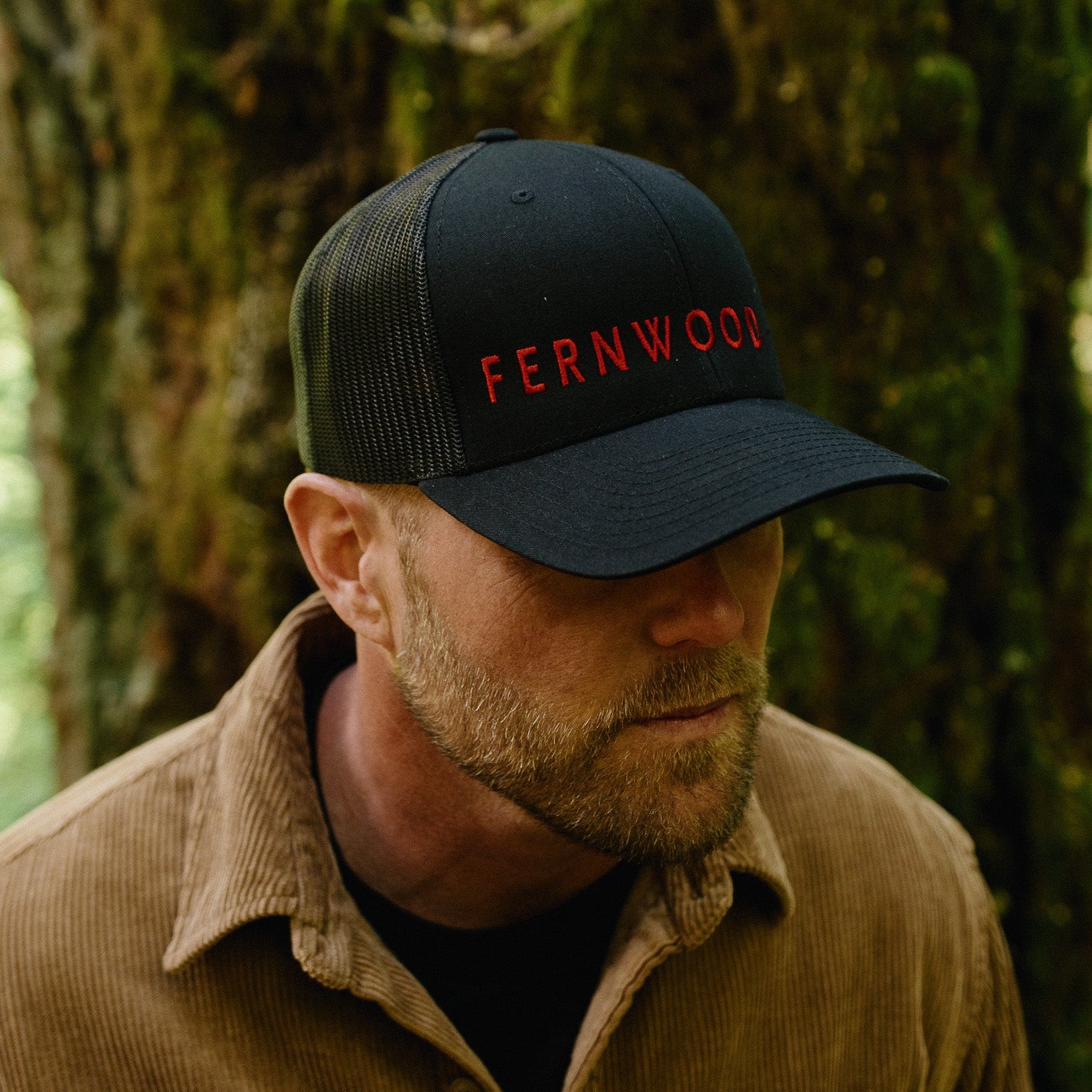 Man wearing a black cap with 'FERNWOOD' in red text, standing in a forest.