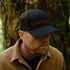 Man wearing a black cap with 'FERNWOOD' in red text, standing in a forest.
