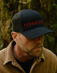 Man wearing a black cap with 'FERNWOOD' in red text, standing in a forest.