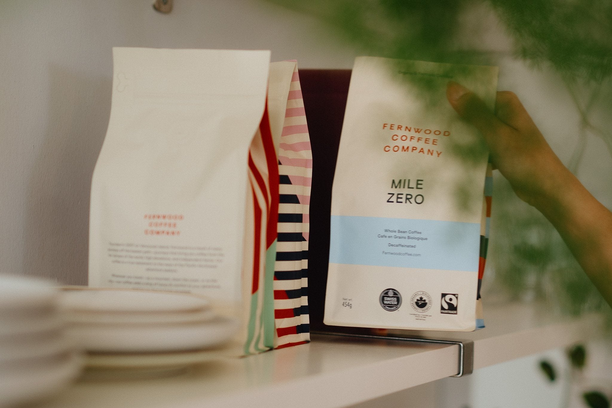 A bag of Fernwood Mile Zero decaf on a shelf beside two striped Fernwood coffee bags, a hand reaching in