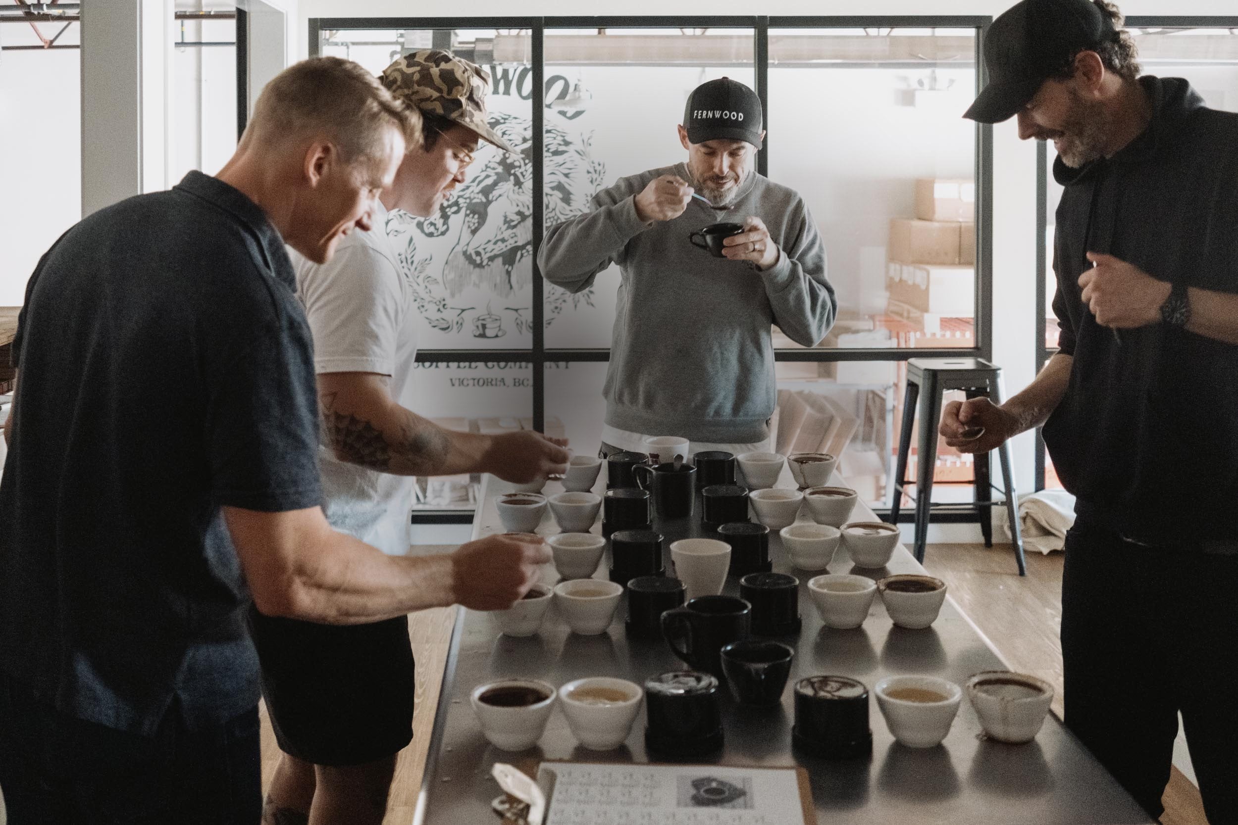 Four Fernwood staff tasting coffee around a steel cupping table lined with dozens of filled cups
