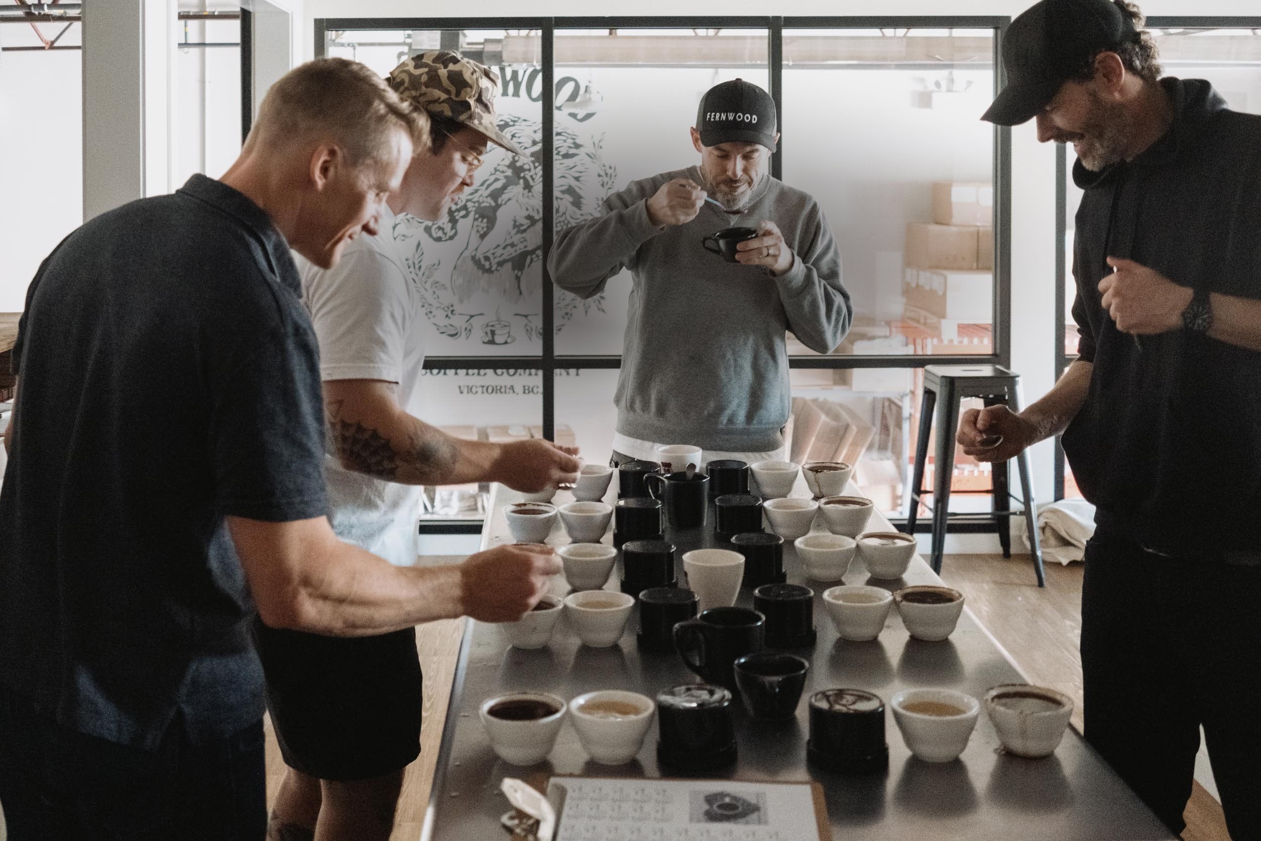 Four Fernwood staff tasting coffee around a steel cupping table lined with dozens of filled cups