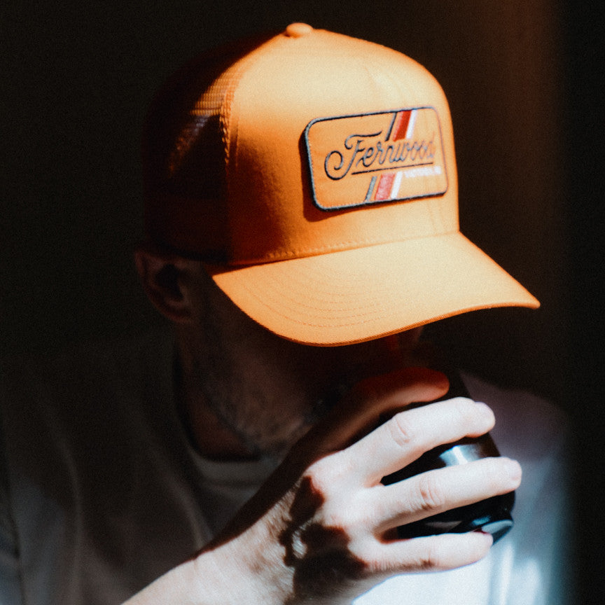 Person wearing an orange cap with a logo, holding a black object against a dark background