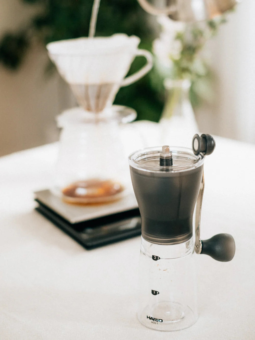 Clear coffee grinder on a white surface, with a blurred background of a pour over coffee maker filter and scale.