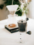 Clear coffee grinder on a white surface, with a blurred background of a pour over coffee maker filter and scale.