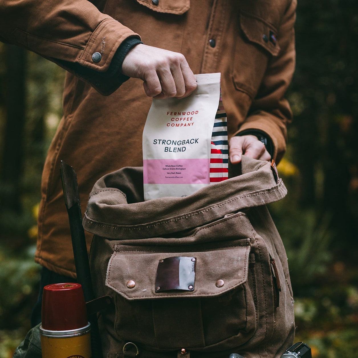 A person pulling a bag of Strongback Blend Coffee from their camping backpack outdoors in a forest.