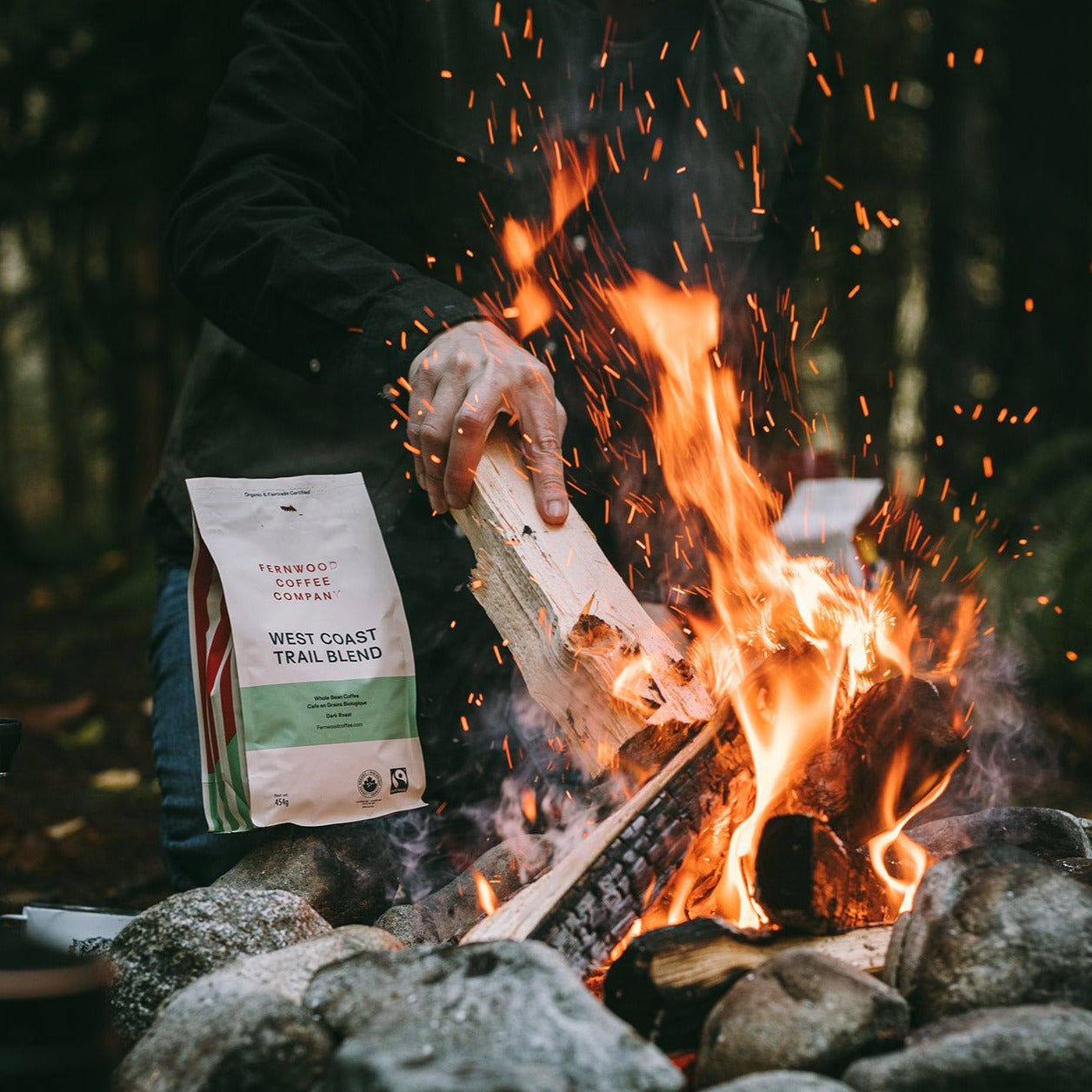 A person poking a log on a campfire outdoors with a bag of West Coast Trail coffee on a nearby rock.