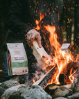 A person poking a log on a campfire outdoors with a bag of West Coast Trail coffee on a nearby rock.