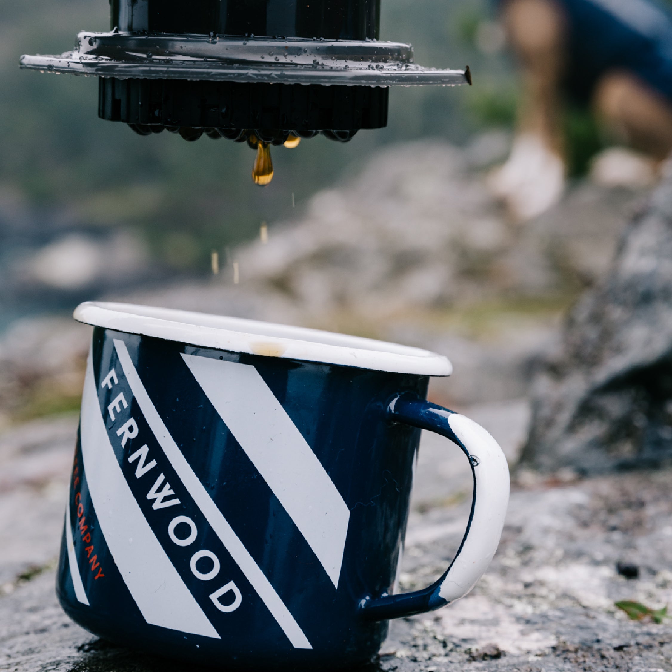 Coffee dripping from the bottom of an Aeropress brewer into a mug outdoors while camping.