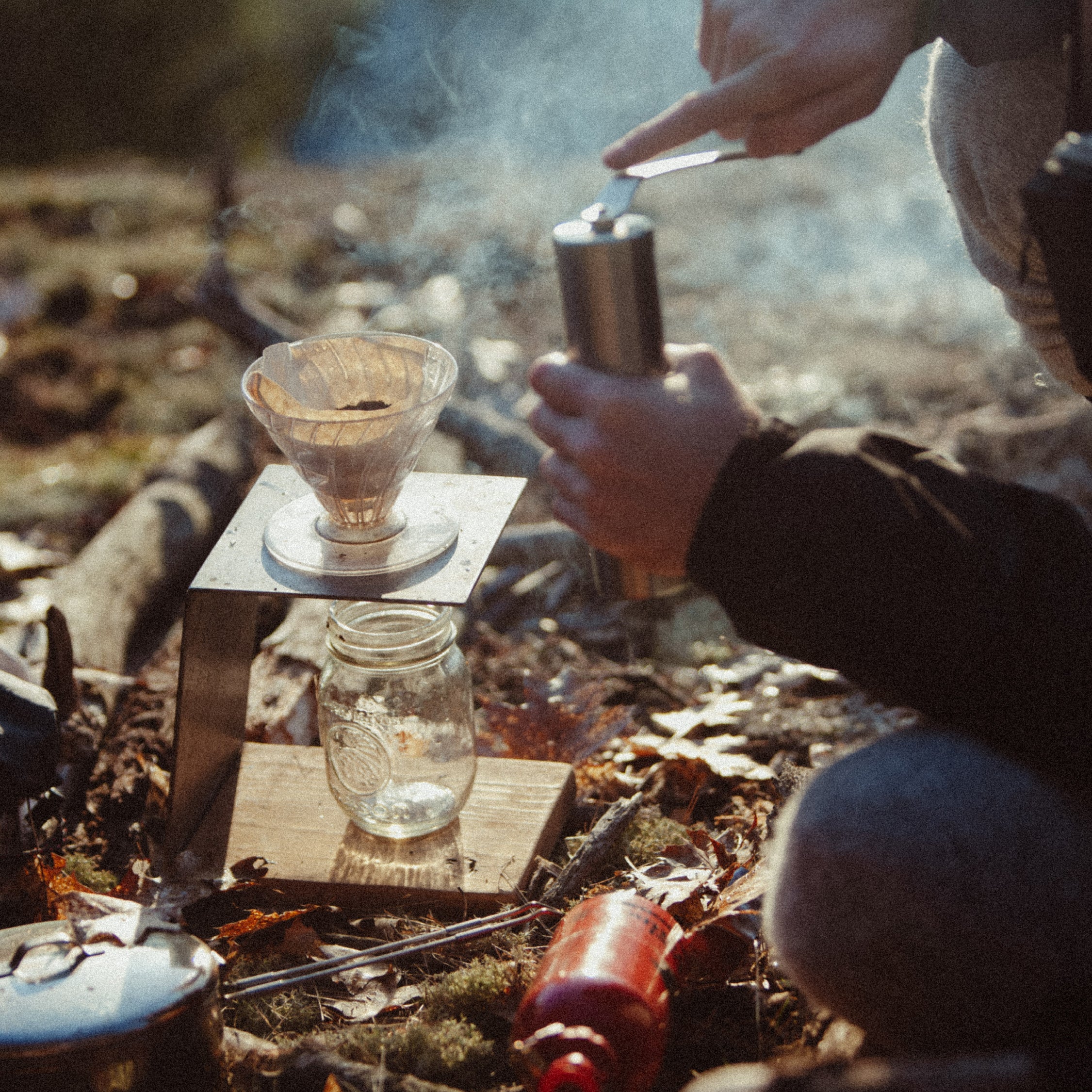 Brewing pour-over coffee outdoors with a hand grinder, mason jar, and campfire smoke in morning light.
