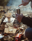 Brewing pour-over coffee outdoors with a hand grinder, mason jar, and campfire smoke in morning light.