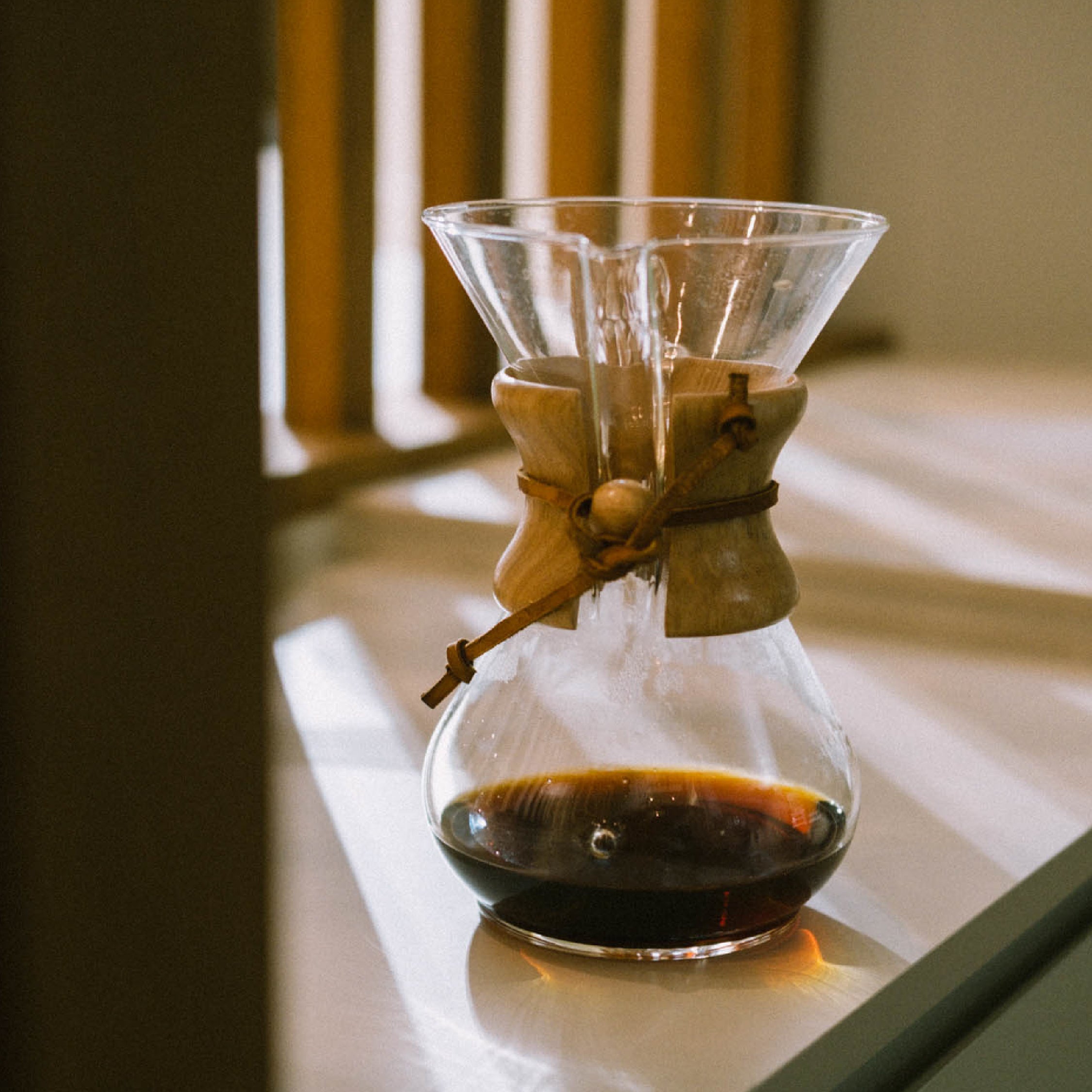 A Chemex coffee brewer sitting in the sunlight on a kitchen counter with some coffee in it.