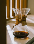 A Chemex coffee brewer sitting in the sunlight on a kitchen counter with some coffee in it.