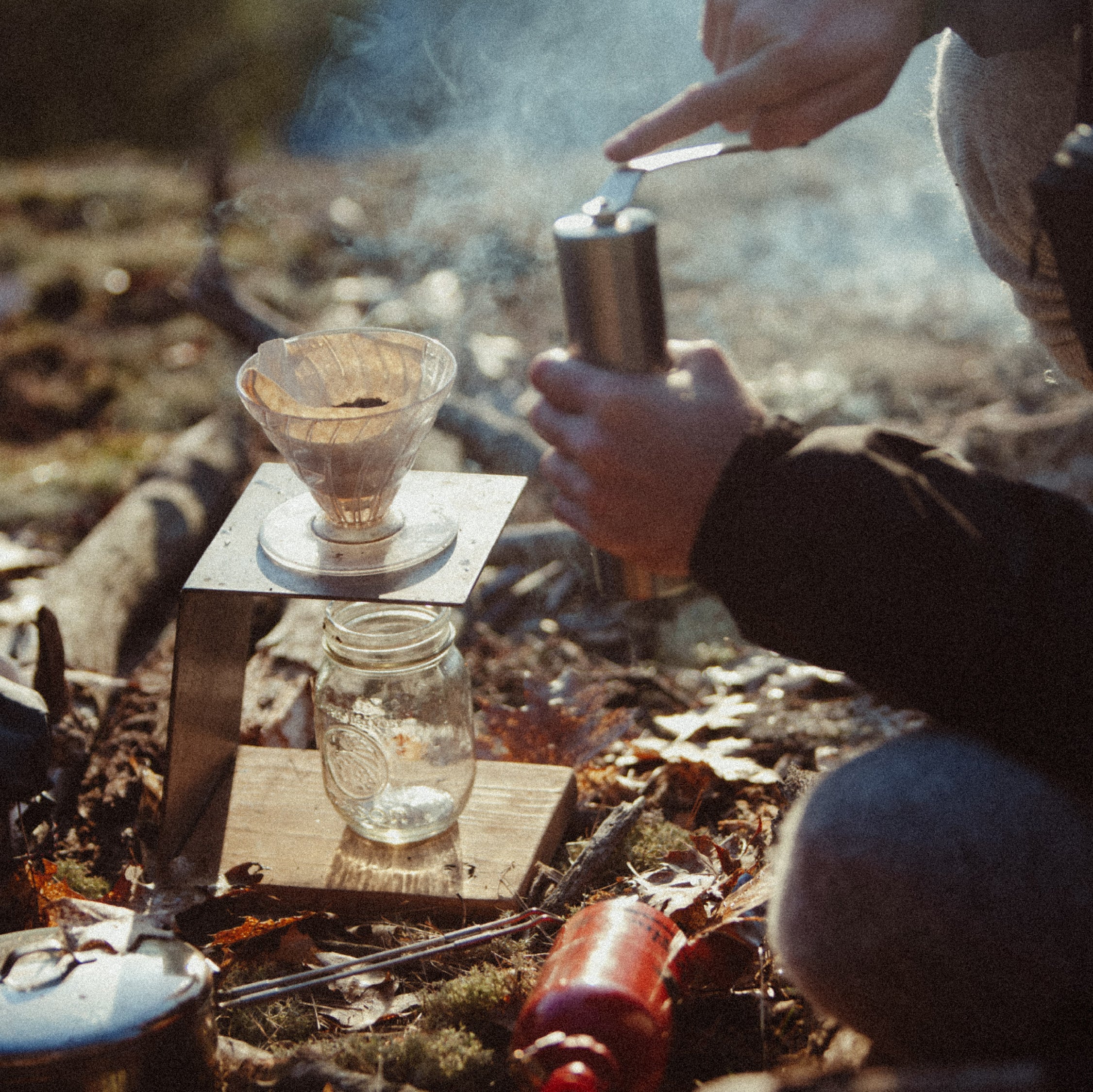 A person manually grinding coffee for a Hario V60 brewer while outdoors camping in the morning light