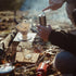 A person manually grinding coffee for a Hario V60 brewer while outdoors camping in the morning light