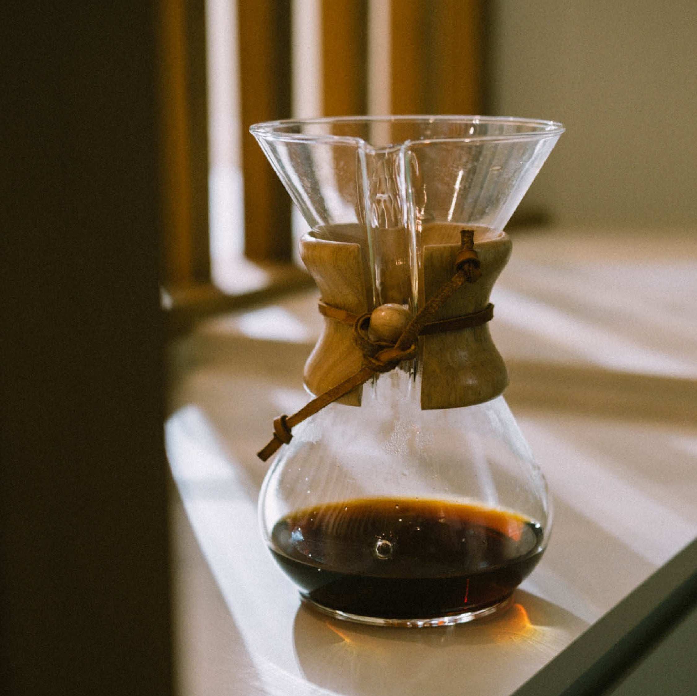 A Chemex coffee brewer sitting in the sunlight on a kitchen counter with some coffee in it.