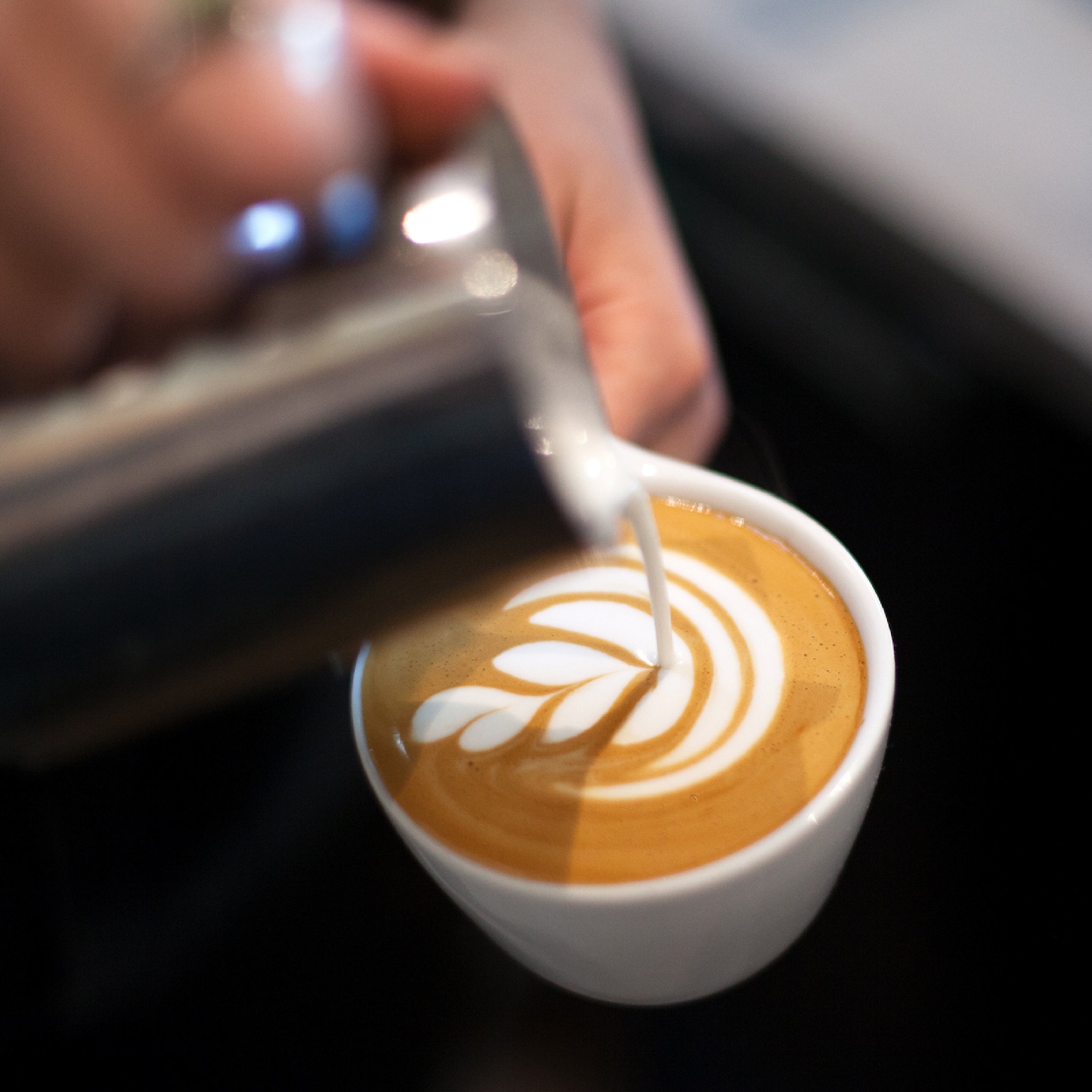 A person pouring milk into a late making latte art on a black counter.