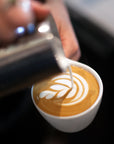 A person pouring milk into a late making latte art on a black counter.